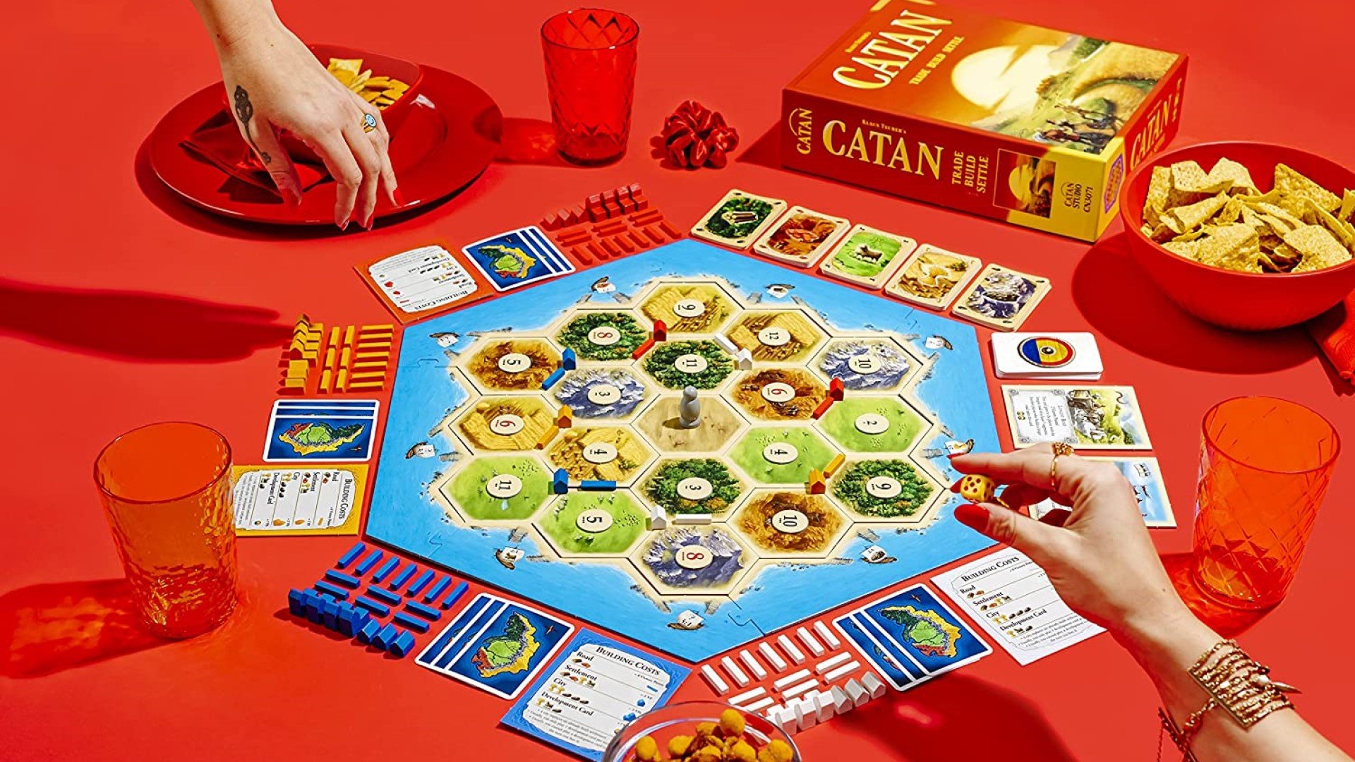 Best classic board games: Catan. Image shows a group of people playing catan with snacks set up around the board on a red background.
