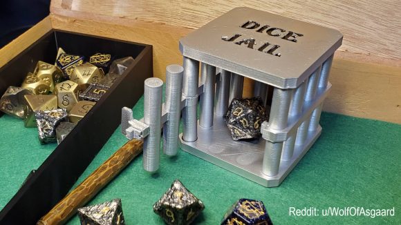 DnD dice DIY torture device - a miniature silver jail with bars and the words 'dice jail' engraved on top. The door is open and a roleplaying die sits inside, with various other dice scattered on the table around the jail.