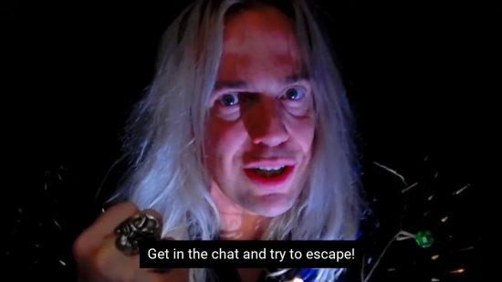 Comedy game twitch stream The Dark Room raising funds for charity - a still from the trailer, John Robertson's face in closeup with the subtitles "Get in the chat and try to escape!"