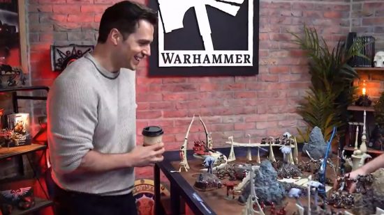 Warhammer 40k Henry Cavill confirmed to star in and executive produce new licensed movies and TV series - Henry Cavill, a well-built man with black hair and wearing a white jumper, holds a coffee cup beside a table of Warhammer figures, in front of a Warhammer logo