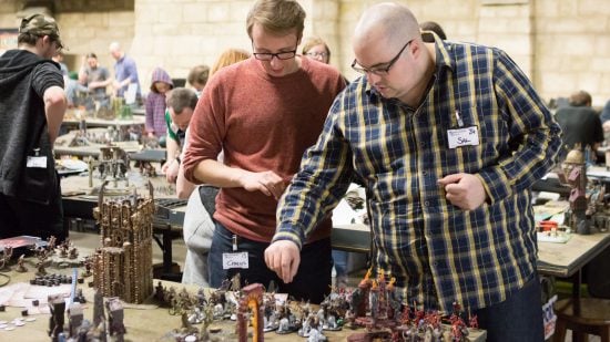What is Warhammer 40k - photograph by Games Workshop of two people playing a tabletop wargame, moving models around a table, rolling dice