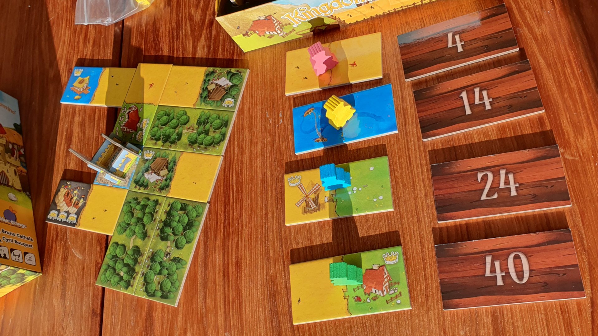 Kingdomino review - example of gameplay, to one side a partially complete kingdom has been built from landscape domino tiles around a castle standee, to the right four different coloured meeples have been placed onto landscape domino tiles to claim them