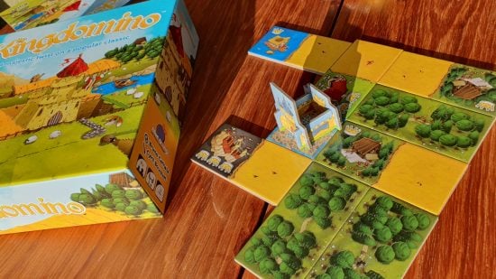 Kingdomino review - example of a partially completed kingdom built from landscape domino tiles around a cardboard castle standee, to the left the colourful game box