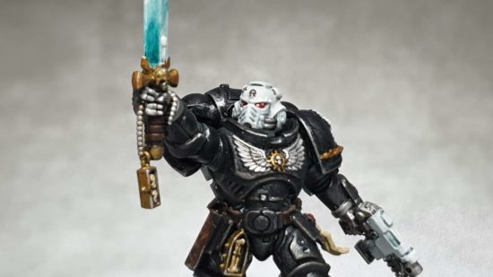 Warhammer 40k Iron Hands lieutenant, a black armoured Space Marine with a white helmet holds aloft a power sword - both his hands are silver, replaced with augmetics