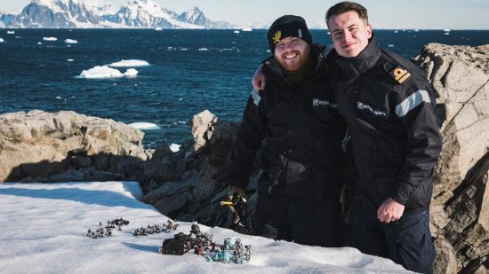 Warhammer 40k competitive player Jonny Talbot and friend Max Friswell, both in black navy uniform, stand in the Antarctic - behind them is deep blue ocean, ice mountains, and in front of them a game of Warhammer 40k played on snow