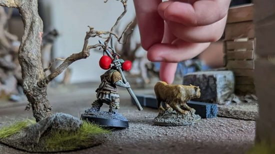 NonCombatTabletop games are like Warhammer without the war - photo by Hydra of Applepicking28, a player's hand reaches into the battlefield beside a miniature man and his dog