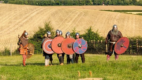Gord game in a real Saxon village longhouse - author photo showing Saxon era battle reenactors in armor, demonstrating a shield wall formation on a sunny day, with farmer's fields on rolling hills in the background