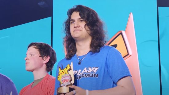 Pokemon TCG North American International Championships final: Cyrus Davis, a white woman with curly brown hair and facial piercings wearing a blue Play Pokemon tshirt, accepts her first-place Pikachu trophy on stage, with her competitor behind her.