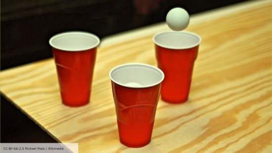 Beer pong rules - a white ball flies into one of three red cups on a wooden table - photo by Michael Mata, hosted CC-BY-SA-2.0 on Wikimedia