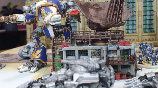 Legions Imperialis review - a warhound Titan looms over a building, preparing to target Legion astartes Kratos heavy tanks