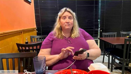 DnD adventure design pioneer Jennell Jaquays, a middle-aged woman with blond hair, wearing a purple shirt, holding a mobile phone, sits in a restaurant - photograph by her wife Rebecca Heineman
