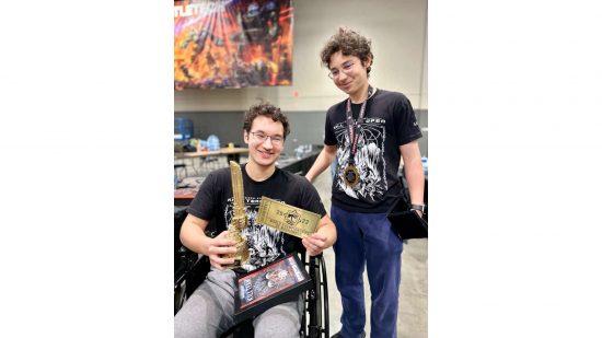 Leander Garrett in a wheelchair, with a golden ticket to the World Championships of Warhammer, and chainsword trophy, beside his brother, at Adepticon 2023 - credit Marc Garrett