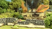 A wrecked tank burns, while an infantryman hiding behind a stone wall aims a bazooka