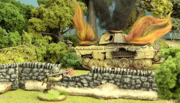 A wrecked tank burns, while an infantryman hiding behind a stone wall aims a bazooka