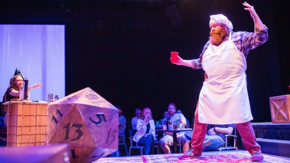 DnD Edinburgh Festival Interactive DnD show - a chef, a giant D20 die, and another actor sat behind a table seen on stage